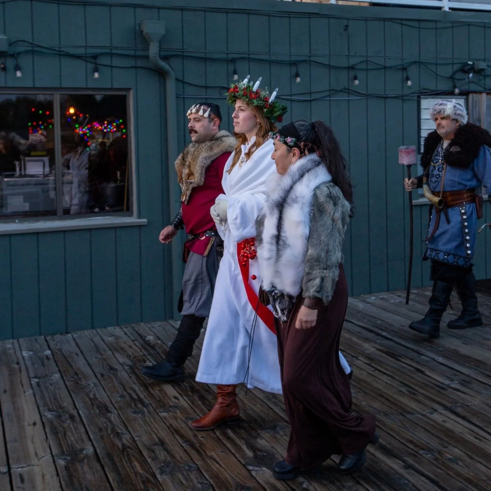 People dressed in medieval or fantasy costumes, participating in a parade or event outdoor on a wooden deck in front of a building with string lights and a window with colorful lights.