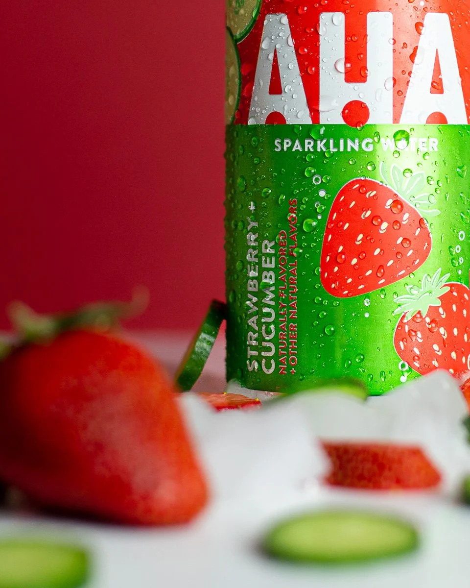 A can of strawberry sparkling water with condensation on the label, surrounded by fresh strawberries and green cucumber slices on a white surface, set against a pink background.