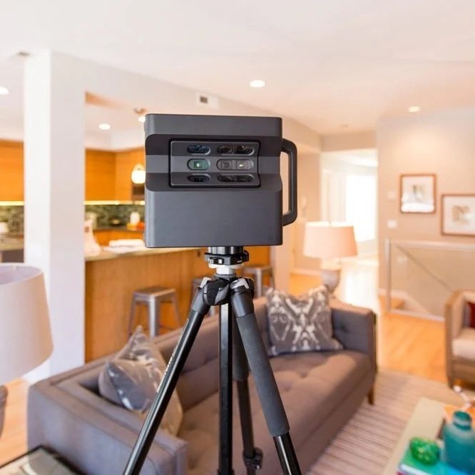 A camera mounted on a tripod inside a living room, with couches, lamps, and framed pictures in the background.