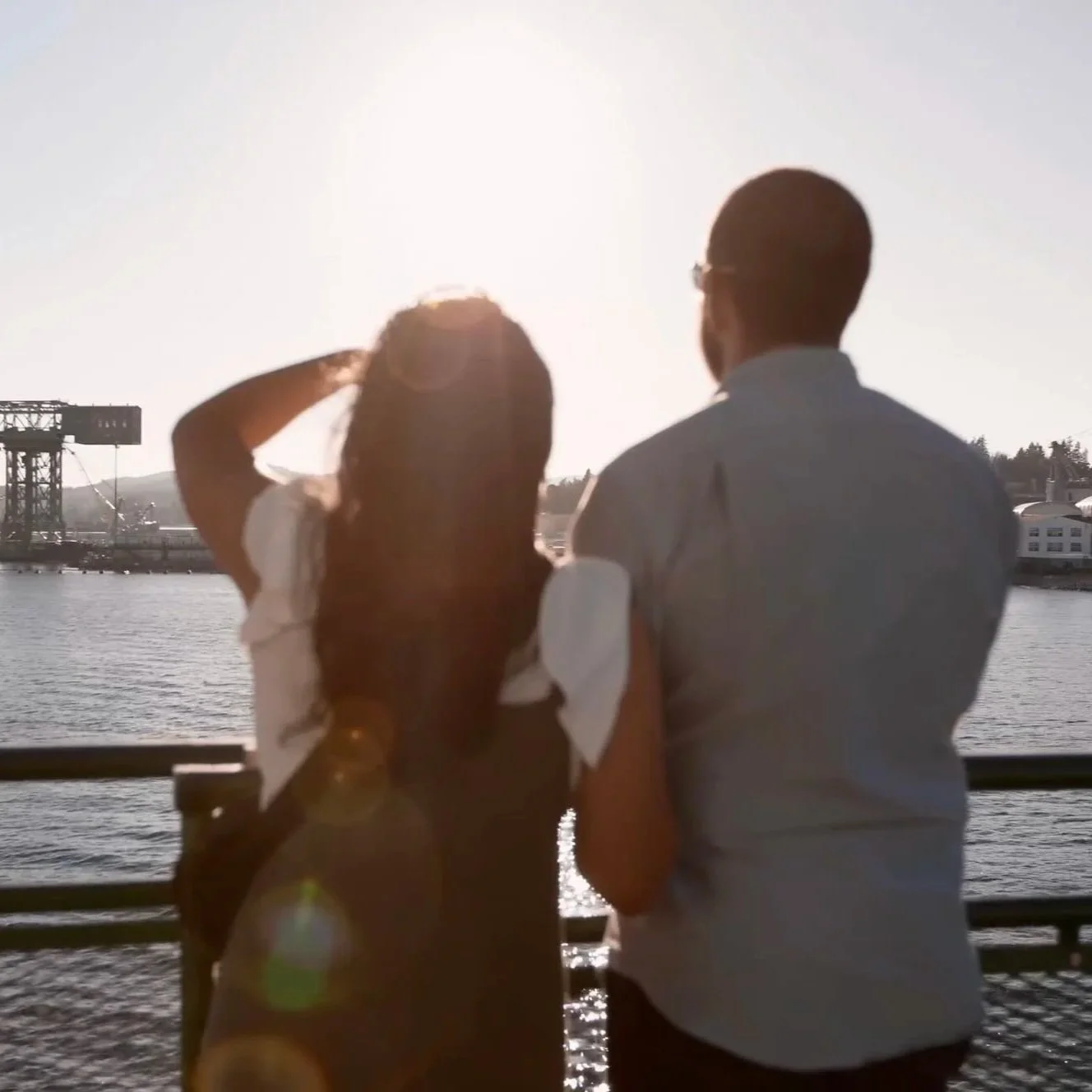 A man and woman standing by a railing by the water, facing the sunset.