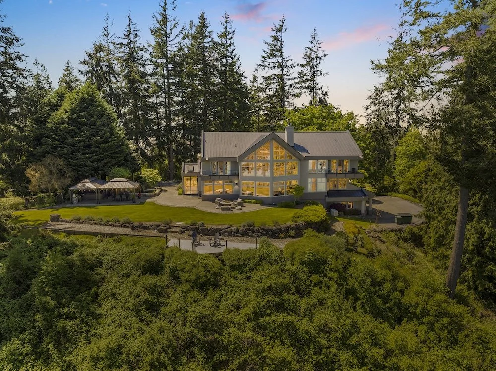 A large modern house with multiple levels and large glass windows, situated in a lush, wooded area during dusk with the interior lights on. There is a patio with outdoor furniture, a small pavilion, and a driveway leading to a garage.