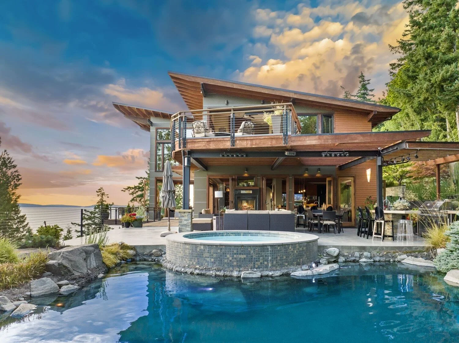 A modern multi-story house with a wooden exterior and large glass windows, an outdoor patio area with seating, a hot tub, and a swimming pool, all set against a scenic backdrop of a lake and trees during sunset.