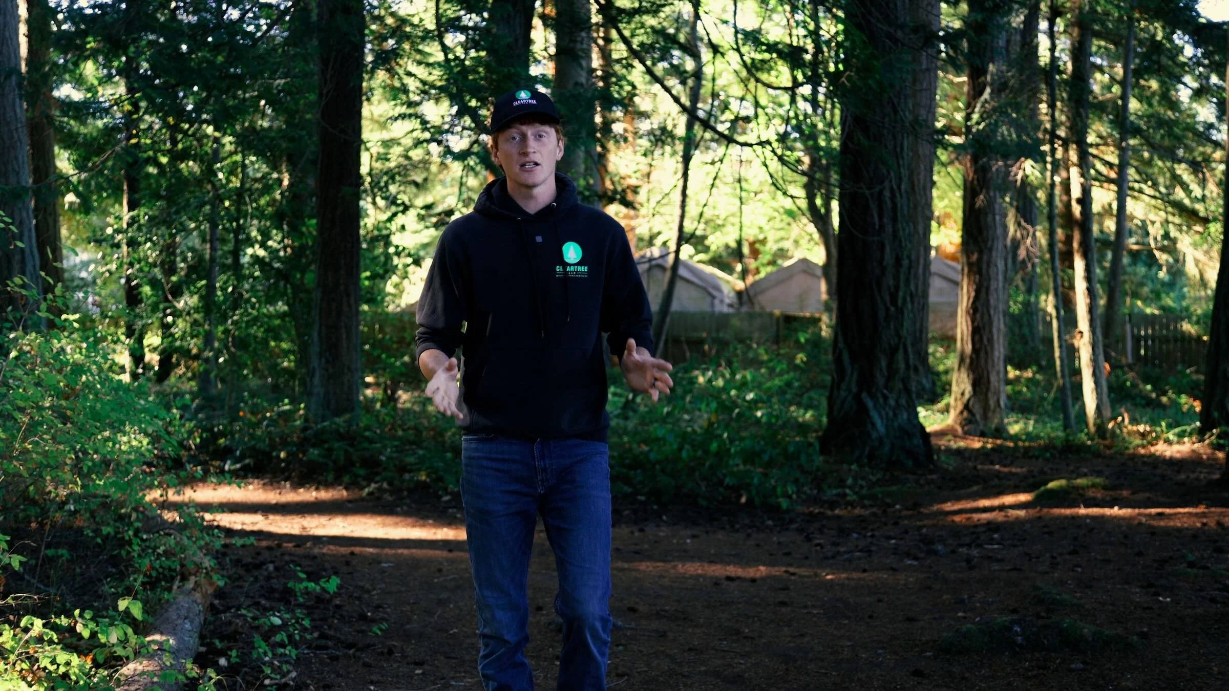 A young man wearing a black hoodie with a logo, jeans, and a cap, walking on a trail through a forest during daytime.