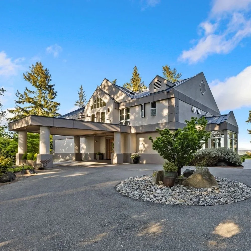 Modern multi-story house with a driveway, surrounded by trees and landscaping, under a blue sky with some clouds.