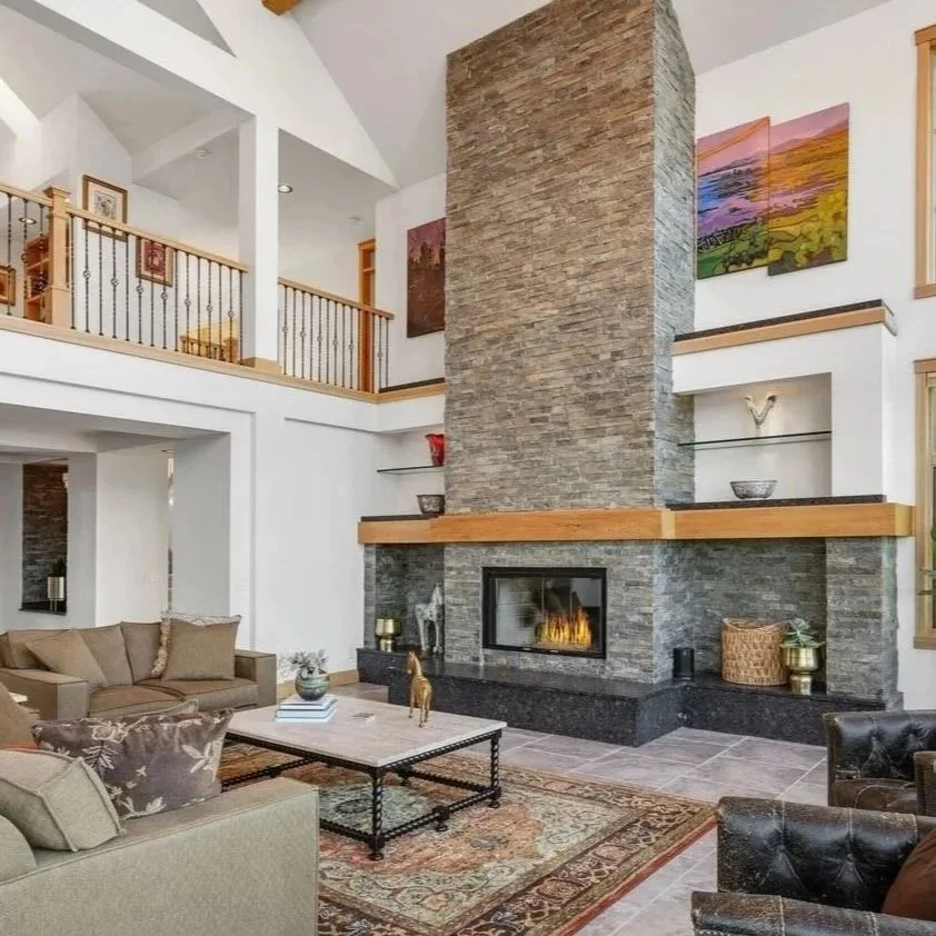 Living room with a large stone fireplace, beige sofas, black leather chairs, and a patterned rug.