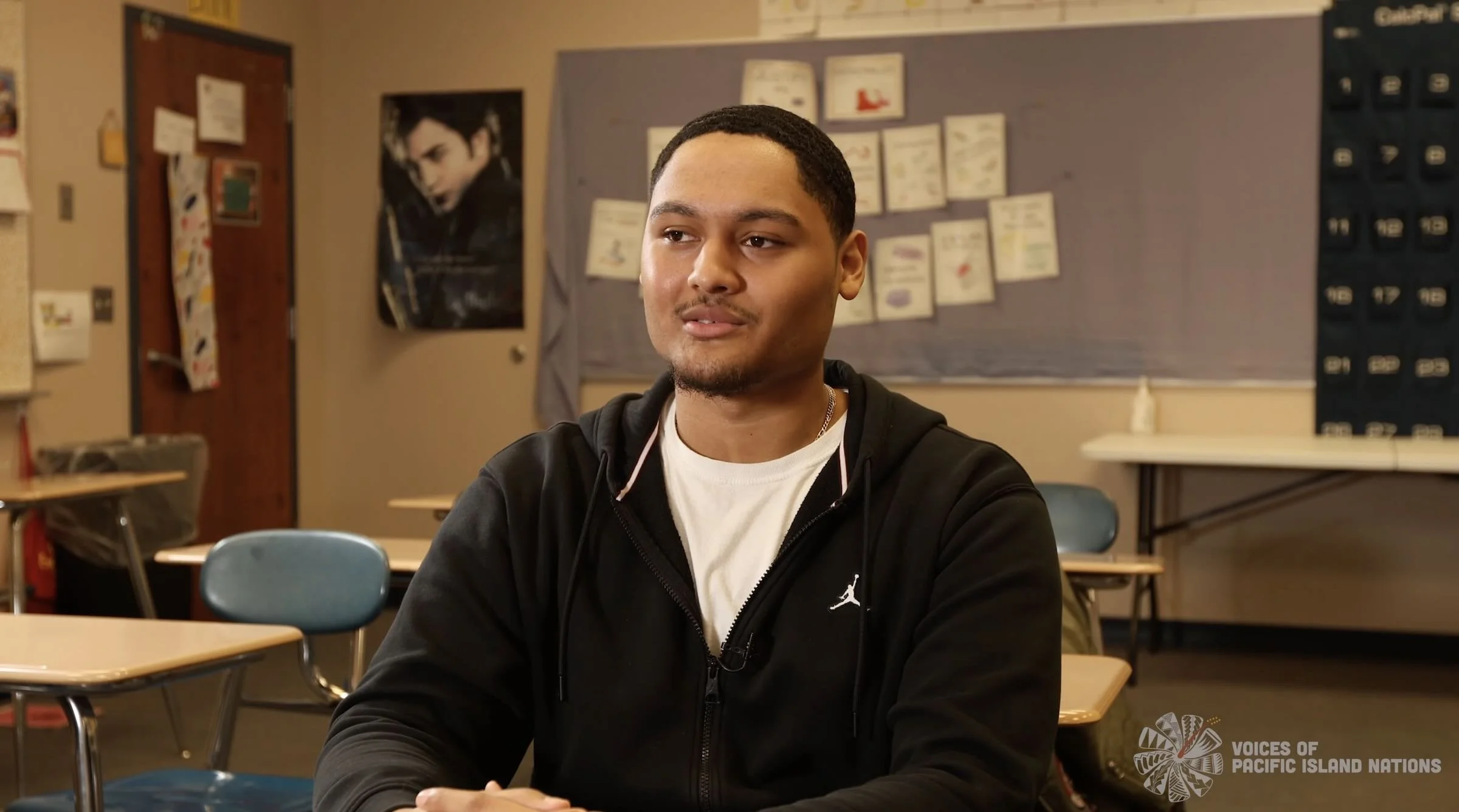 A young man with short black hair and a light mustache, sitting in a classroom, wearing a black zip-up hoodie over a white t-shirt, with a poster and bulletin board in the background.