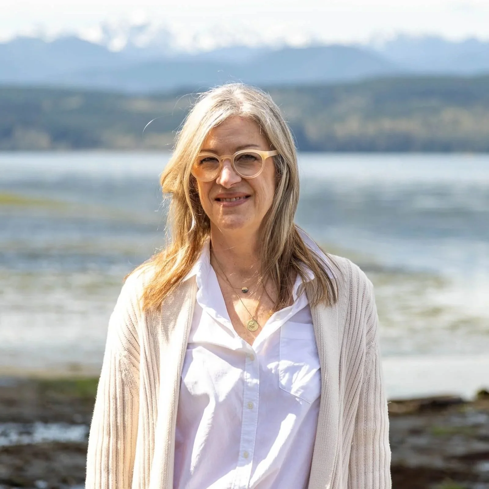 A woman with long blonde hair and glasses standing outdoors near a body of water with mountains in the background.