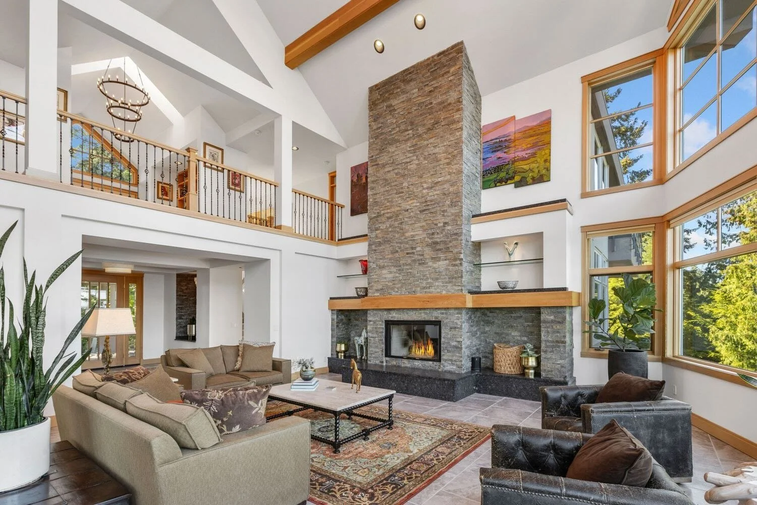 Living room with modern stone fireplace, large windows, comfortable sofa, armchairs, and indoor plants, in a house with high ceilings and wood accents.