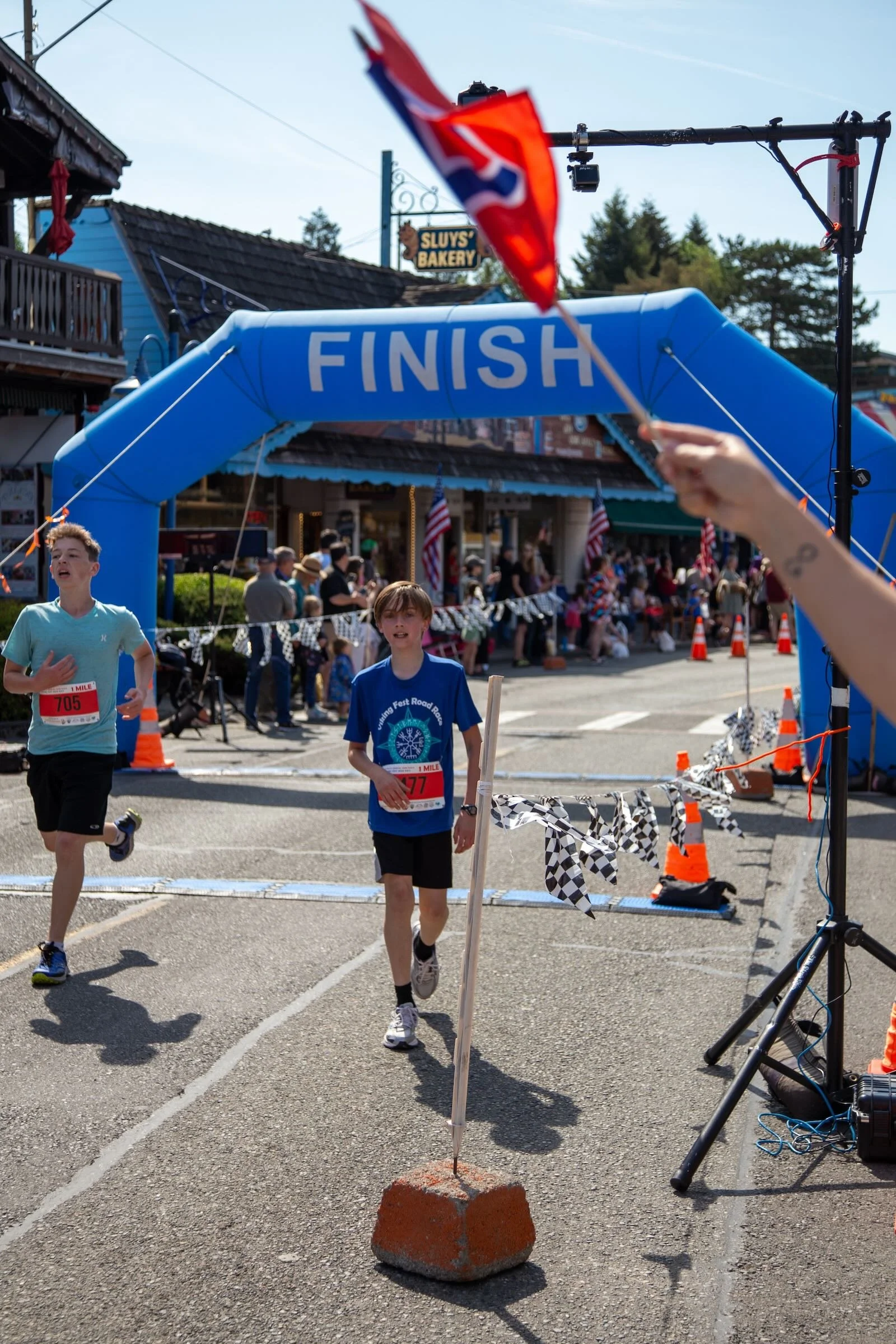 Two boys crossing the finish line of a race under a blue arch with the word 'FINISH'. A person is holding a small flag that is part of the scene. Spectators and storefronts are visible in the background.