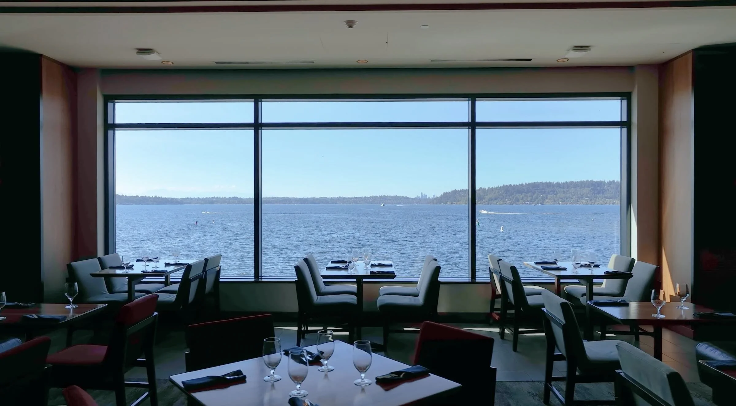 Dining room with large window overlooking a body of water with boats and distant land, set with glasses, napkins, and utensils.