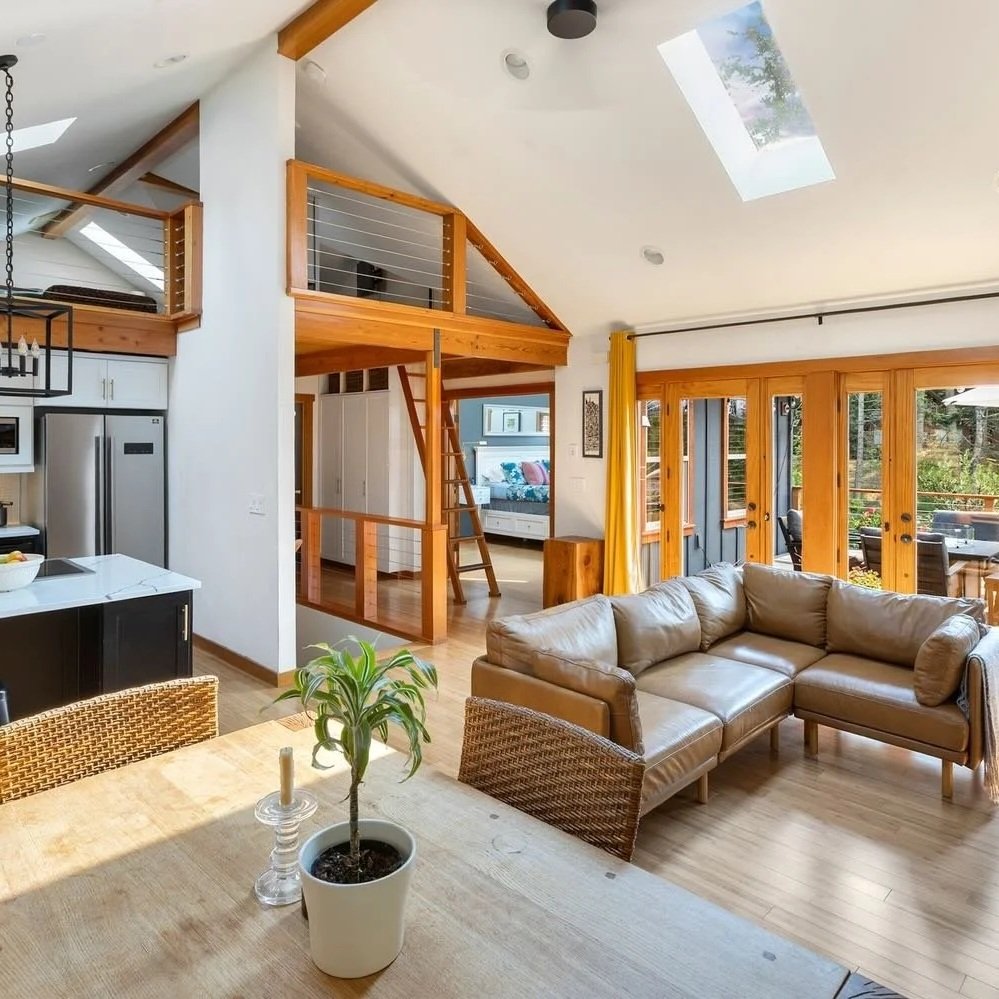 Open-concept living area with a tan sectional sofa, large glass sliding doors with yellow curtains, a skylight, and a kitchen area with black cabinets. A loft with a wooden railing is visible above.