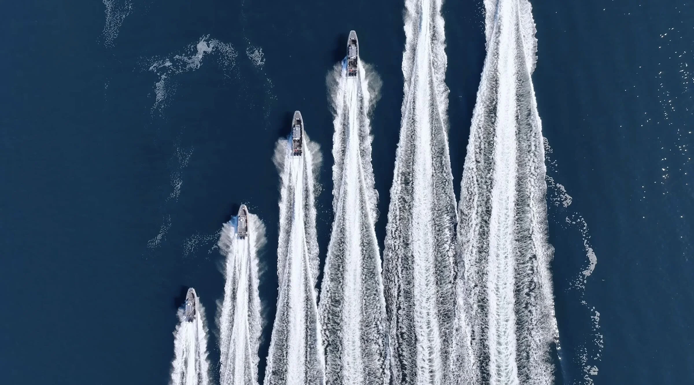 Five speedboats in a formation creating wakes in the water, viewed from above.