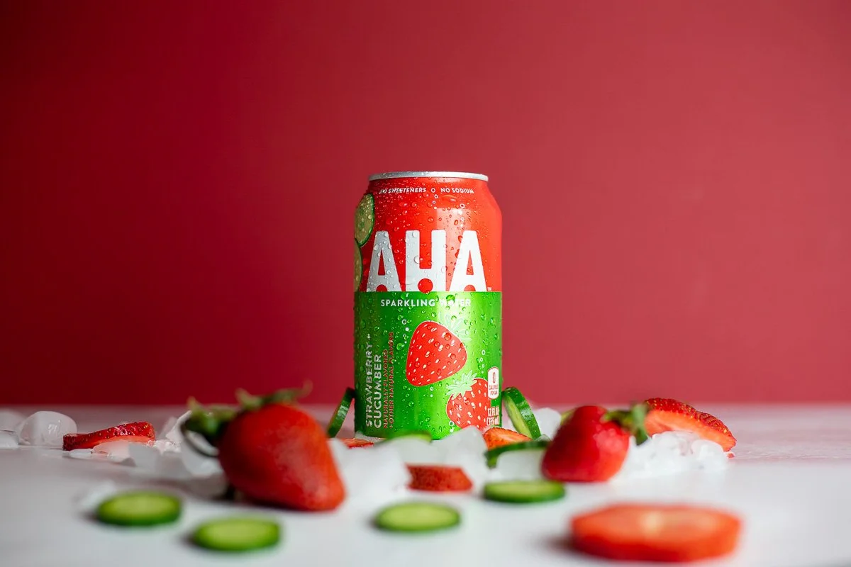A can of AHA sparkling water with strawberry cucumber flavor, surrounded by strawberries, cucumber slices, and ice on a white surface against a red background.