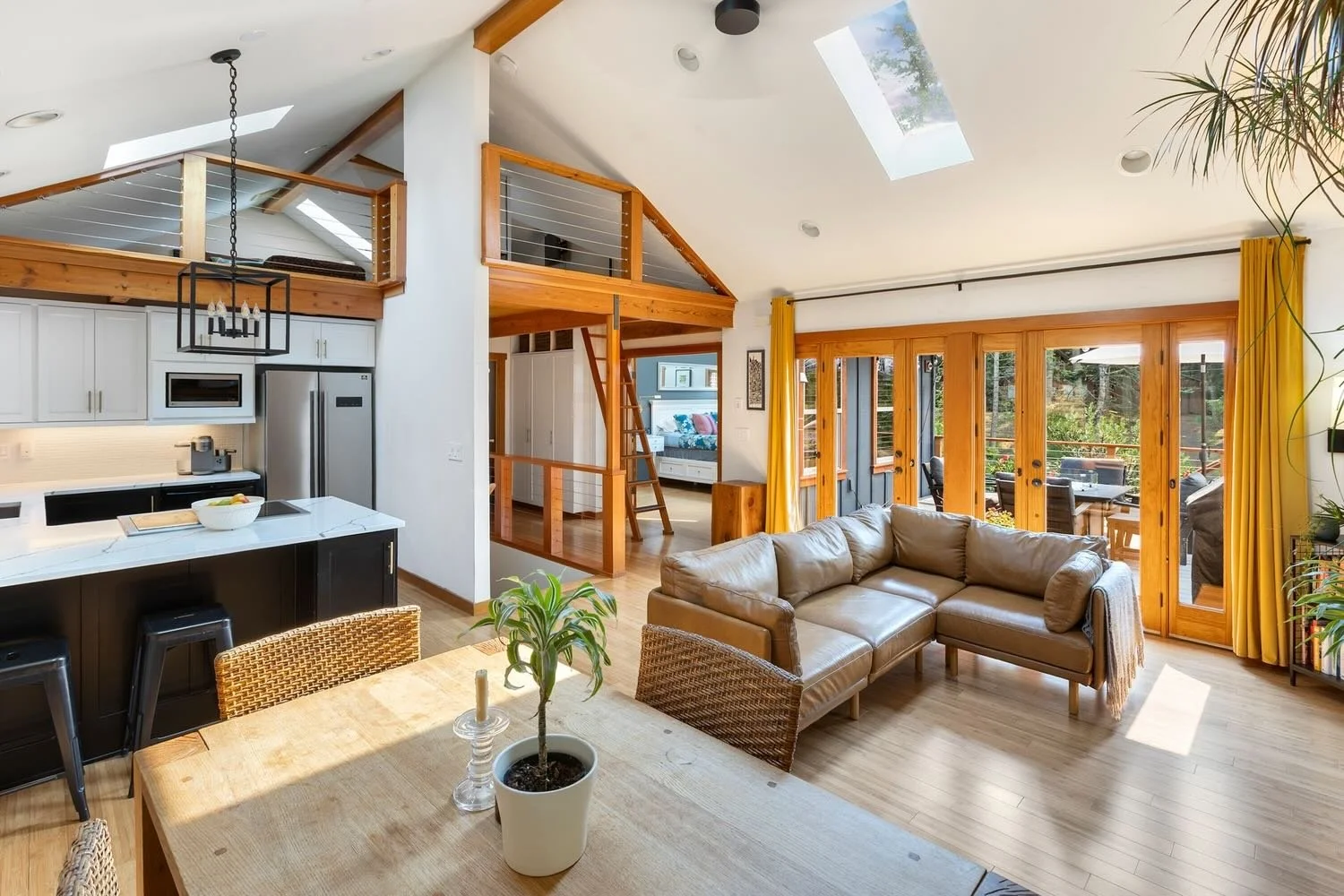 Open-concept living room and kitchen with wooden accents, large sliding glass doors leading to outdoor deck, beige sectional sofa, wooden dining table with plant centerpiece, and bright natural light from skylights.