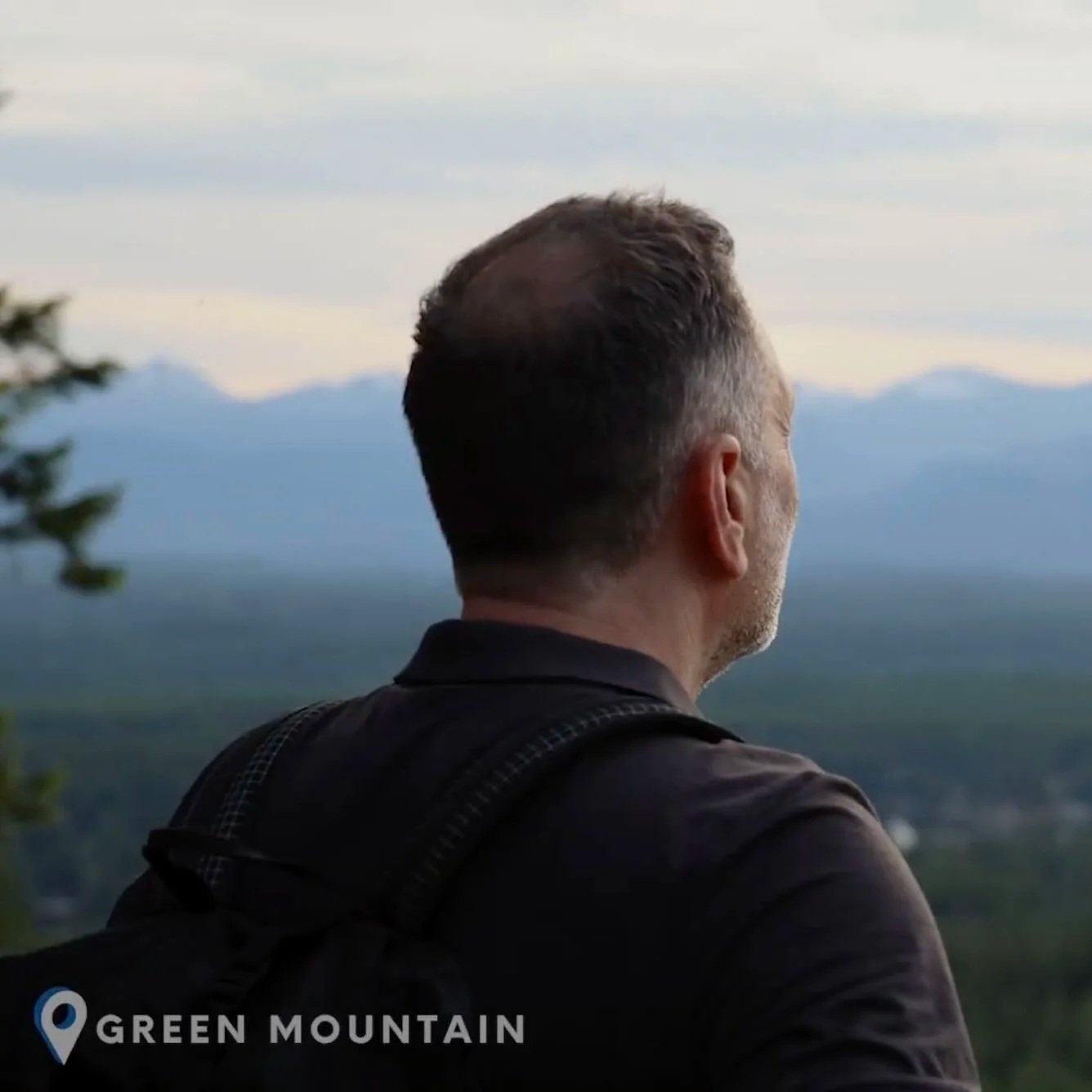 A man with short hair, looking out at a mountain landscape, wearing a black shirt and carrying a backpack, with the label 'Green Mountain' in the bottom left corner.