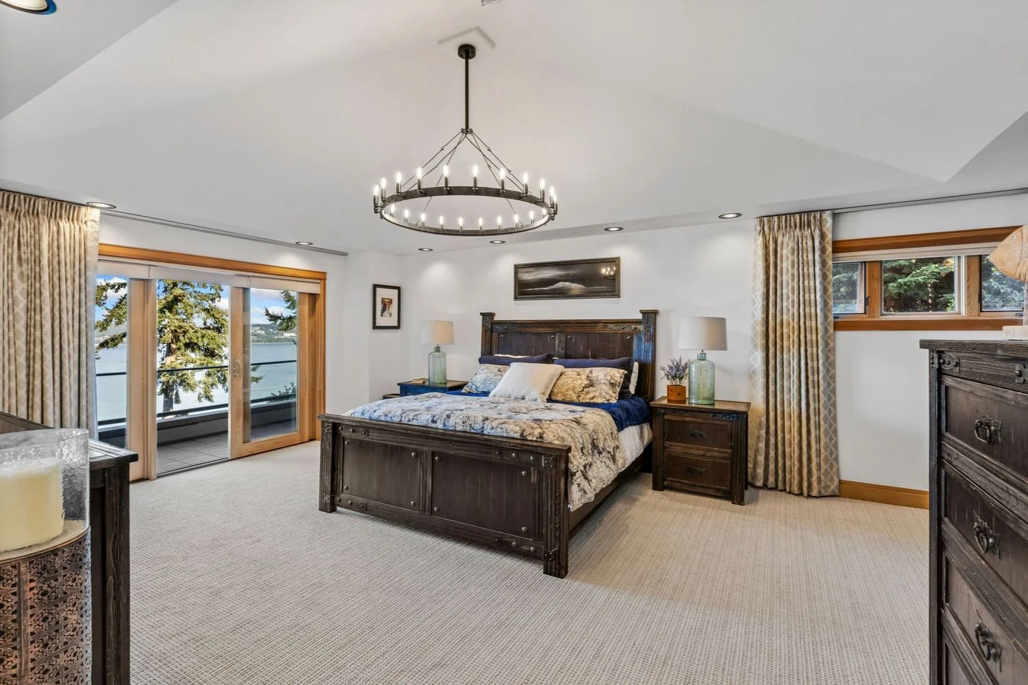 A bedroom with a large wooden bed, nightstands with lamps, and a view of trees and water through sliding glass doors and windows.
