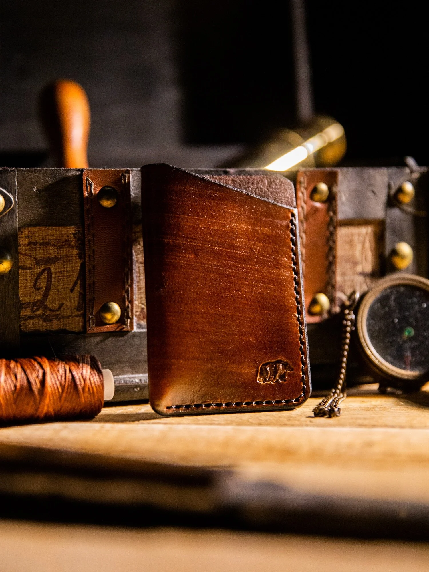 Brown leather wallet with a small logo, placed on a wooden surface, with leather and brass items in the background.