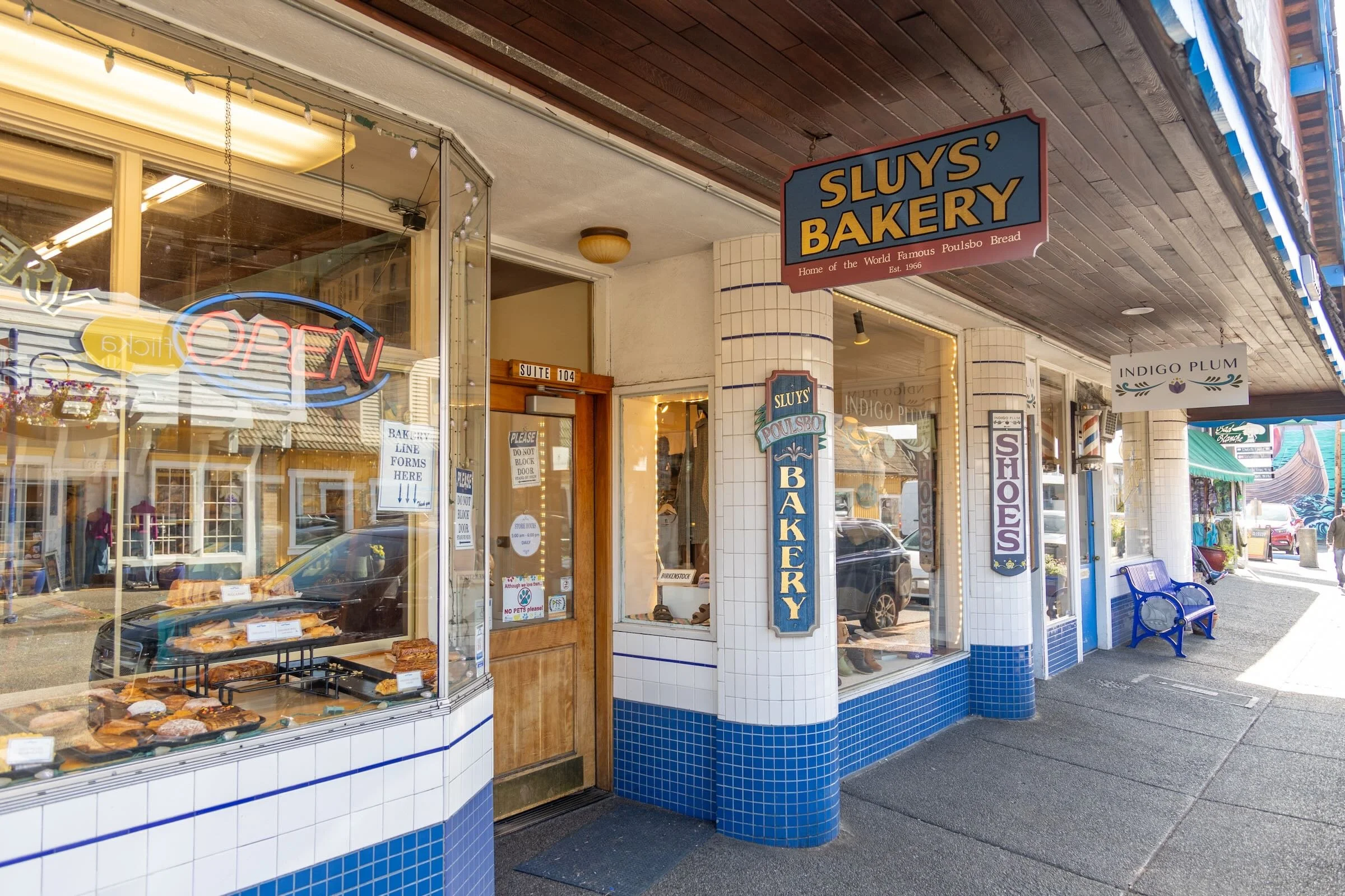 Exterior of Sluy's Bakery with a large window display of baked goods, a blue and white tiled facade, a wooden door, and signs advertising the bakery and shoes outside.