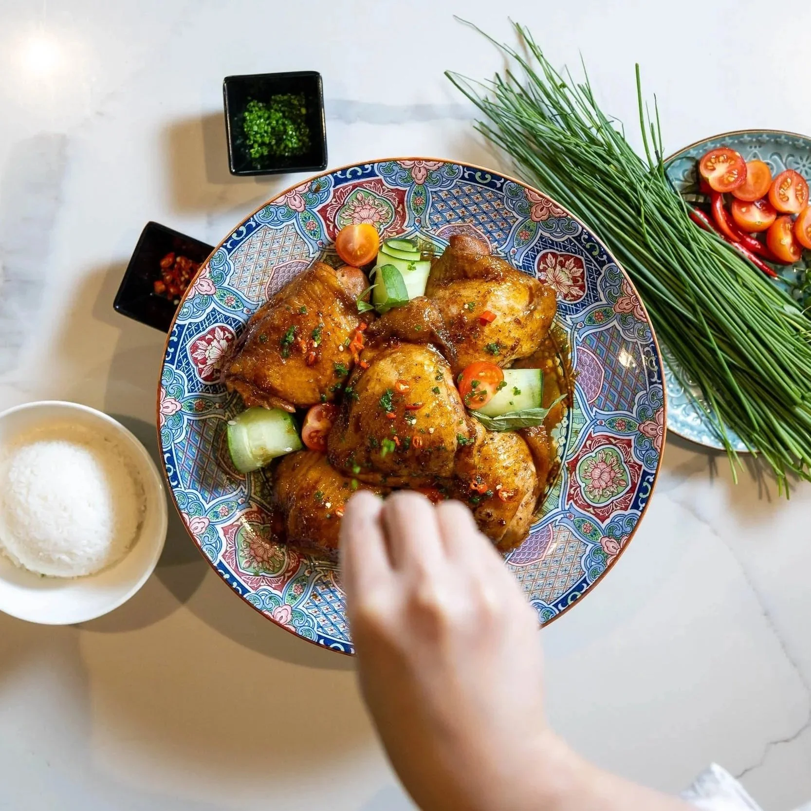 A colorful bowl of glazed chicken drumsticks garnished with chopped herbs and sliced vegetables, with side dishes of chopped tomatoes, chives, and shredded coconut on a light-colored surface.