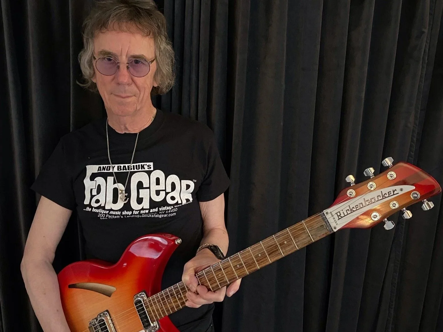 A man with gray hair and purple-tinted glasses holding a red electric guitar. He is wearing a black T-shirt with white text and a necklace, standing against a black curtain background.