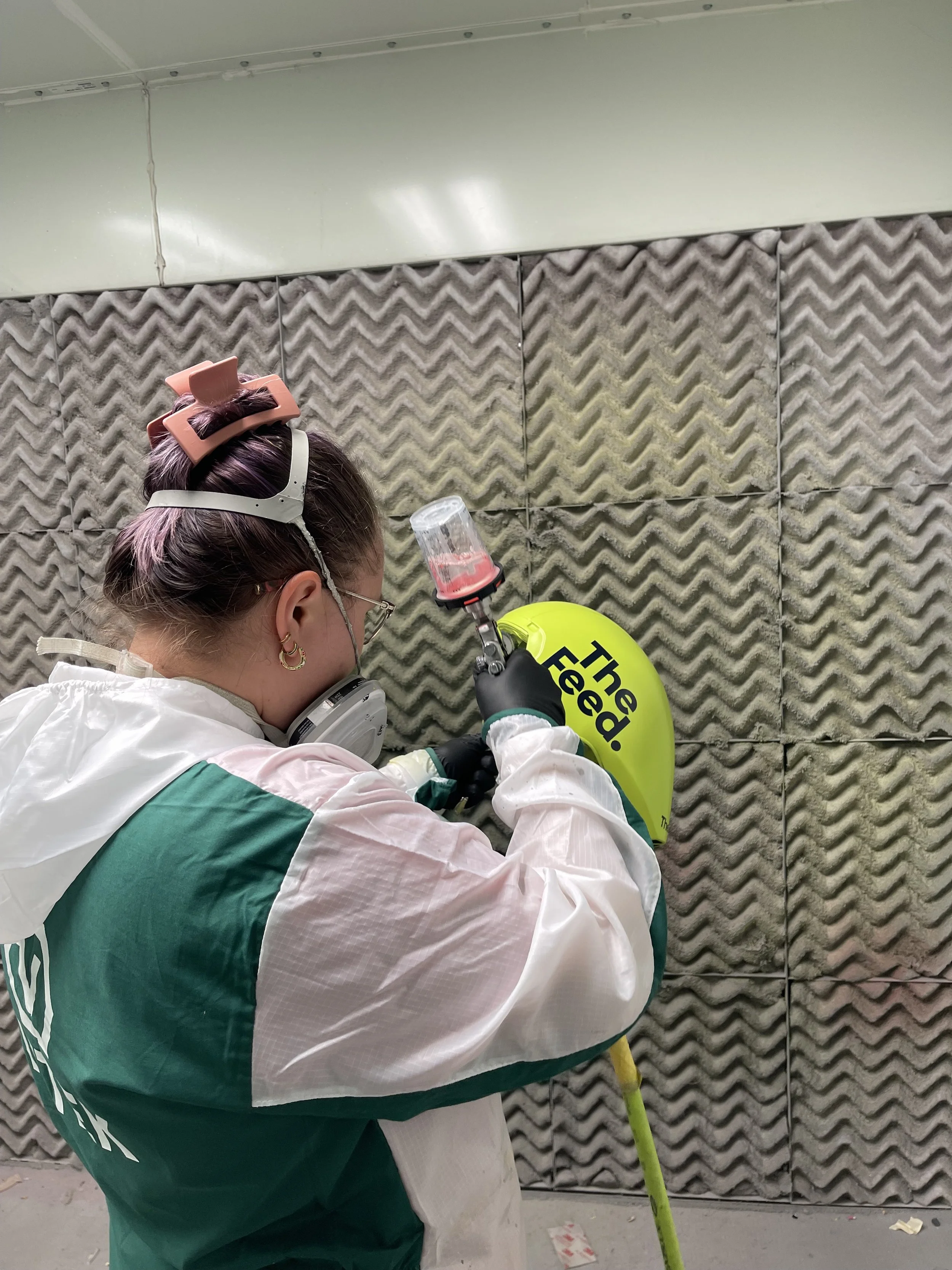 A person in protective gear spray paints a green helmet with the phrase 'The Feud' in black letters in an enclosed soundproof room.