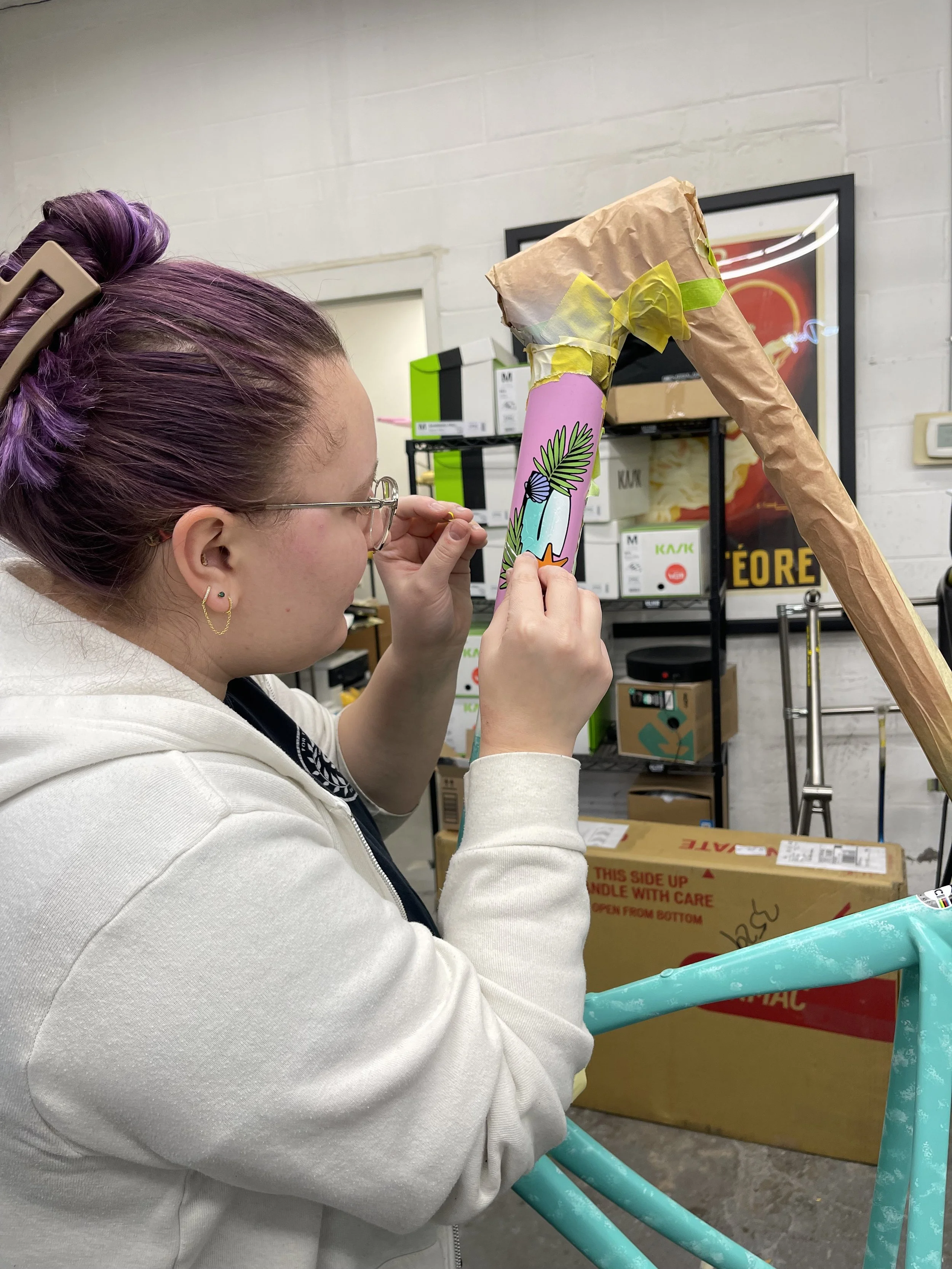 A woman with purple hair and glasses is painting a colorful, cartoon-style design on a cardboard cone, possibly for a creative project or packaging, in a cluttered workspace or storage area.