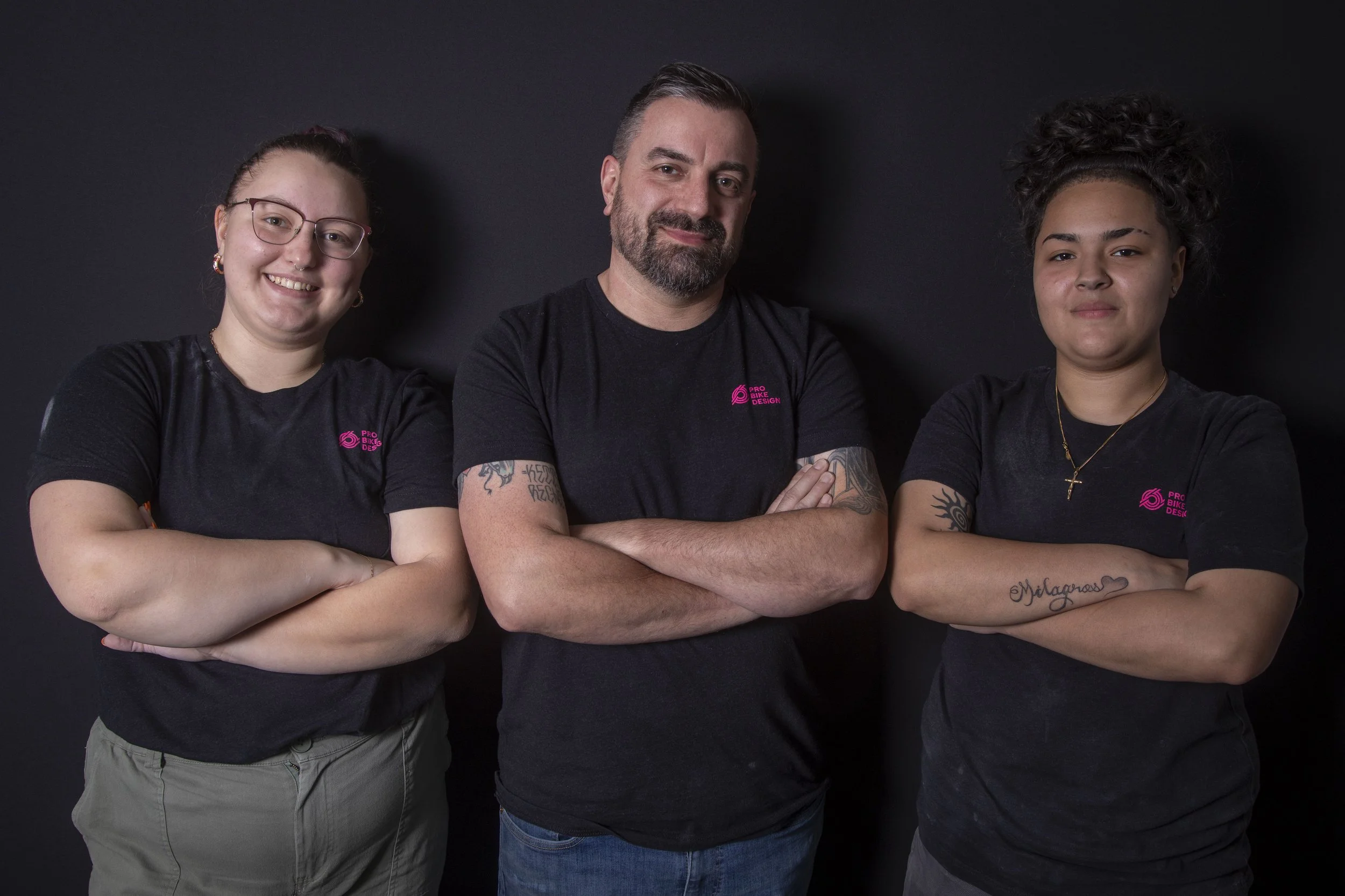 Three people standing against a black background, all wearing black t-shirts with pink logos, arms crossed, smiling confidently.