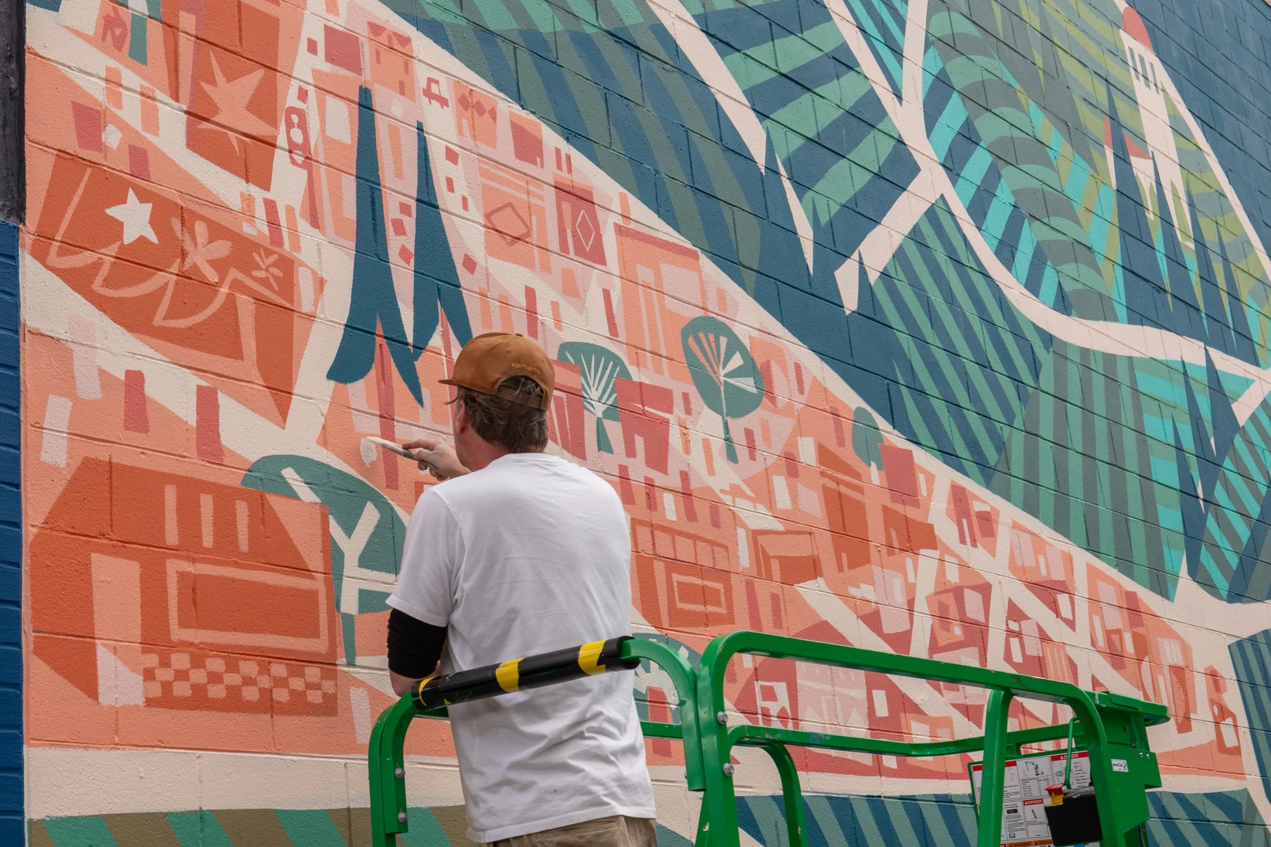 A person painting a colorful mural on a brick wall using a paintbrush, standing on a green lift.