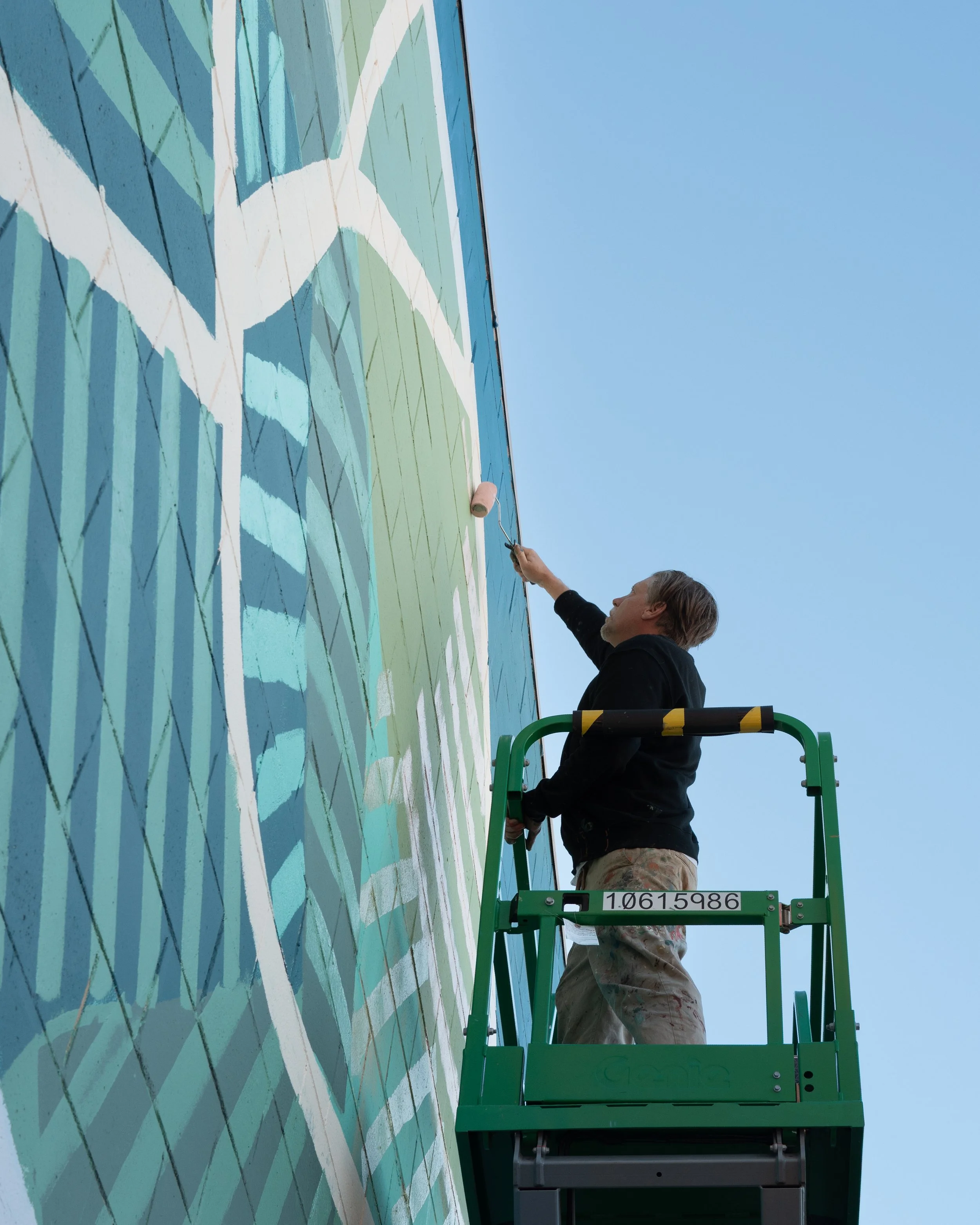 A person on a green lift painting a mural on a tall wall with blue sky in the background.