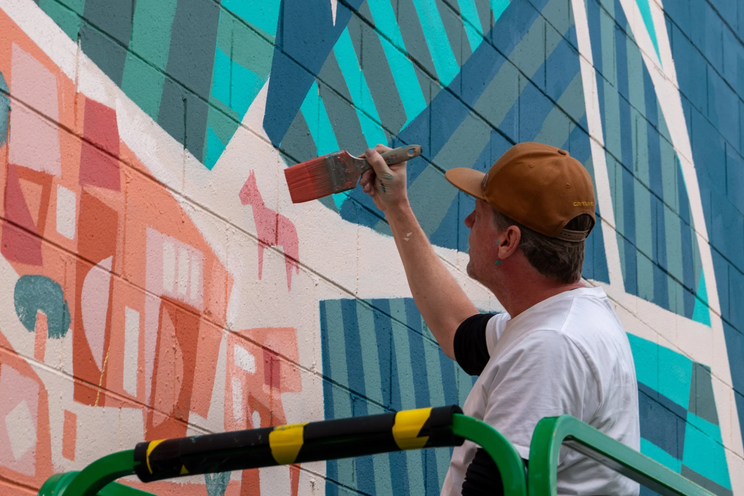 A man wearing a brown cap and white t-shirt painting a colorful mural on a brick wall, standing on a green and yellow scaffold.