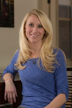 Woman with long blonde hair in a blue shirt sitting indoors, smiling.