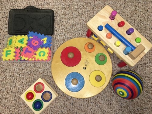 Assorted children's educational toys on a carpet, including a shape puzzle, a box of foam alphabet and number puzzle pieces, a colorful ball, and a wooden pounding bench with a hammer.
