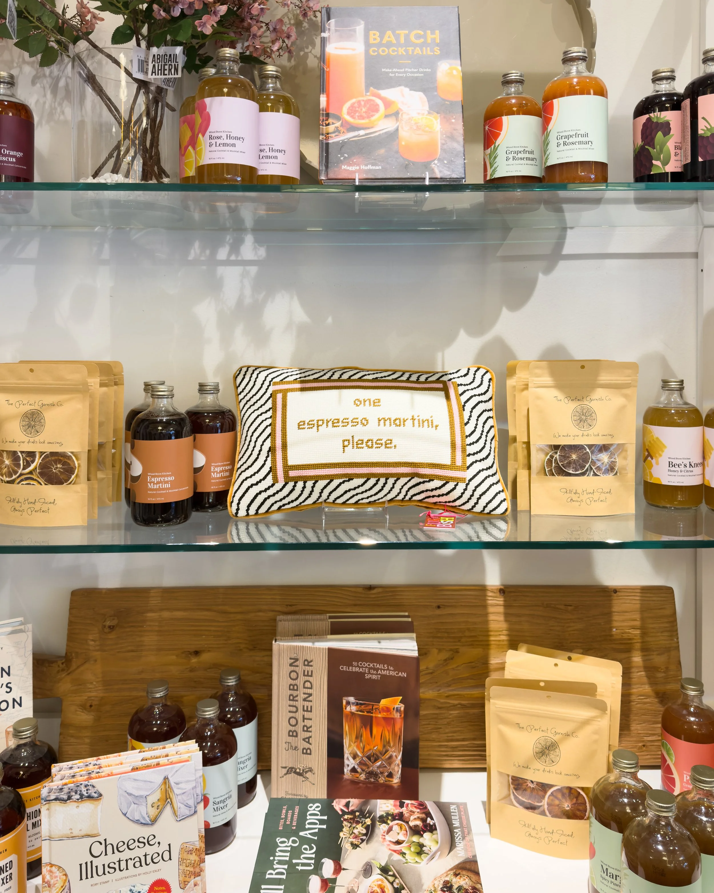 Boutique display of cocktail mixers, garnish kits, cocktail books and a needlepoint pillow reading “one espresso martini, please.”