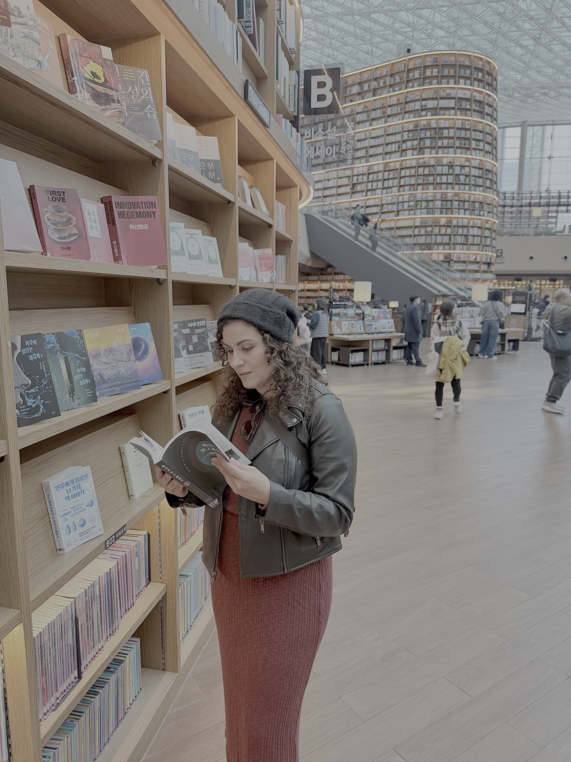 A woman with curly hair wearing a gray beanie and a black leather jacket, reading a book in a bookstore or library with wooden shelves filled with books. There are other people in the background walking around and browsing.
