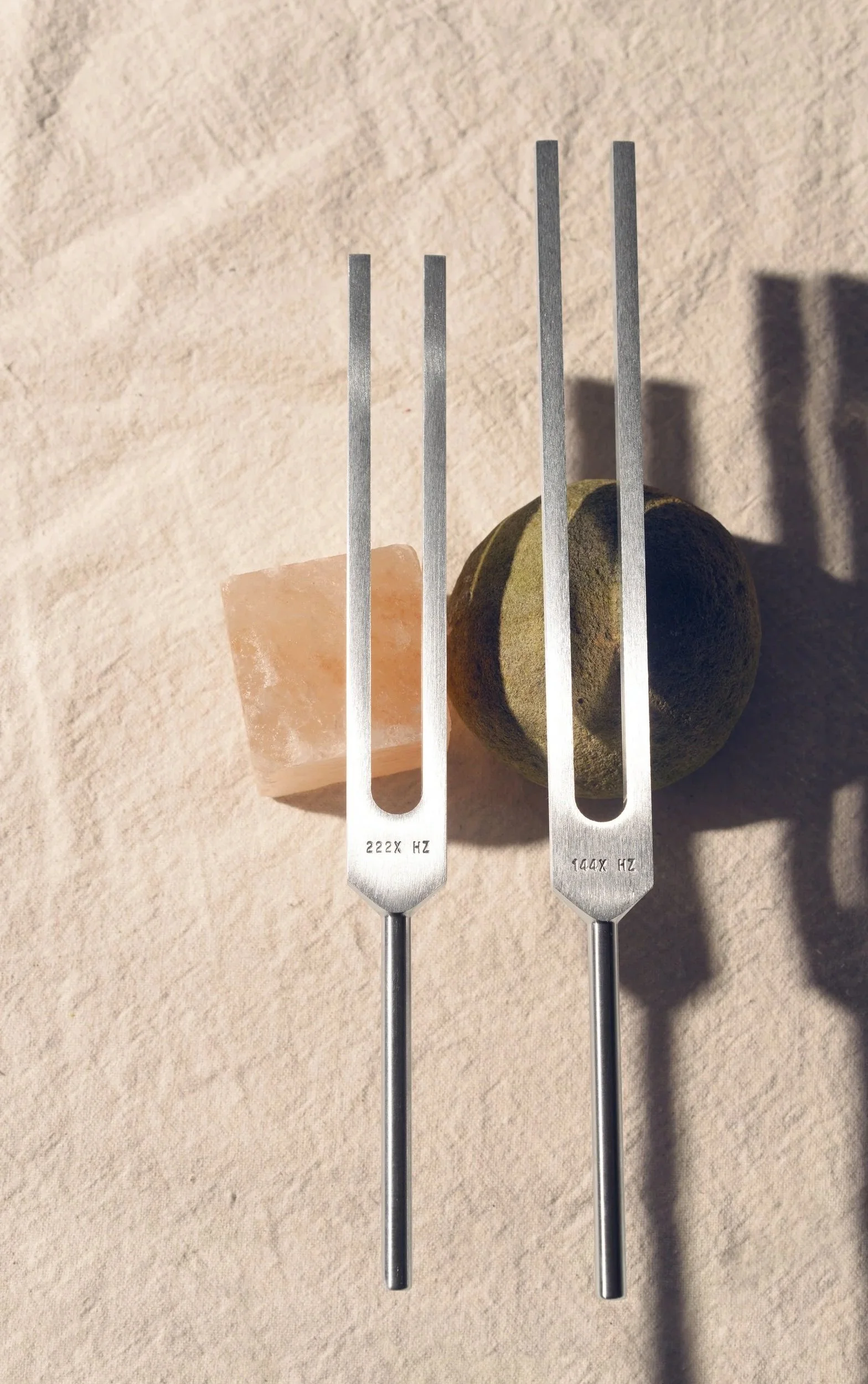Two tuning forks, one marked 222 Hz and the other 144 Hz, placed on a beige surface with a salt rock and a round stone or ball between them.