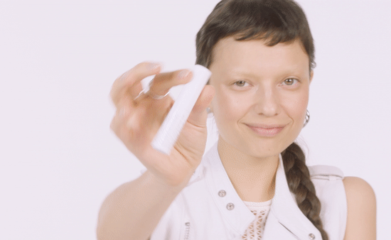 A woman with short brown hair and a braid, smiling, holding a white object in front of her. She is wearing a white vest with silver buttons and a patterned shirt underneath.