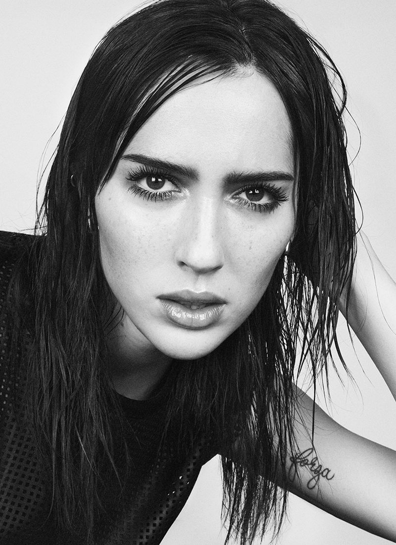 A black and white portrait of a woman with wet, dark hair, looking directly at the camera with intense eyes.