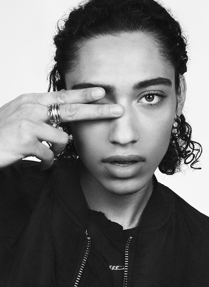 Black and white portrait of a woman with wet hair, holding her hand up to her eye in a peace sign gesture, wearing rings and earrings.