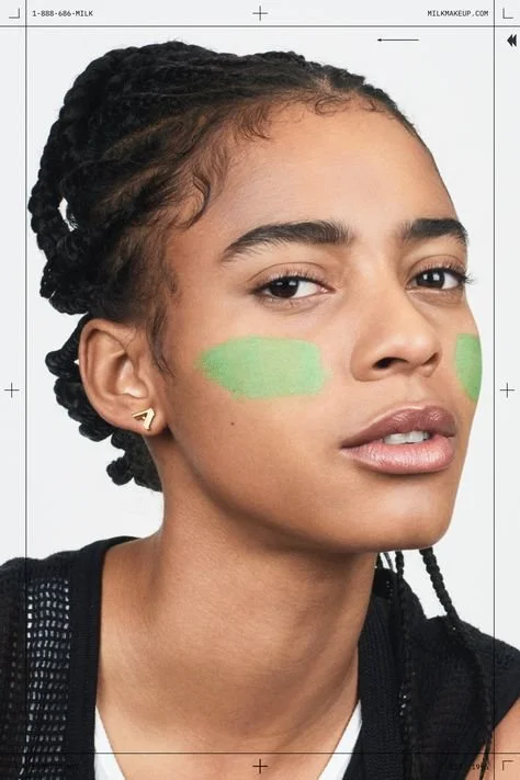 Young woman with partially braided hair has green face paint on her cheeks, wearing a black mesh top and gold earrings, looking at the camera.