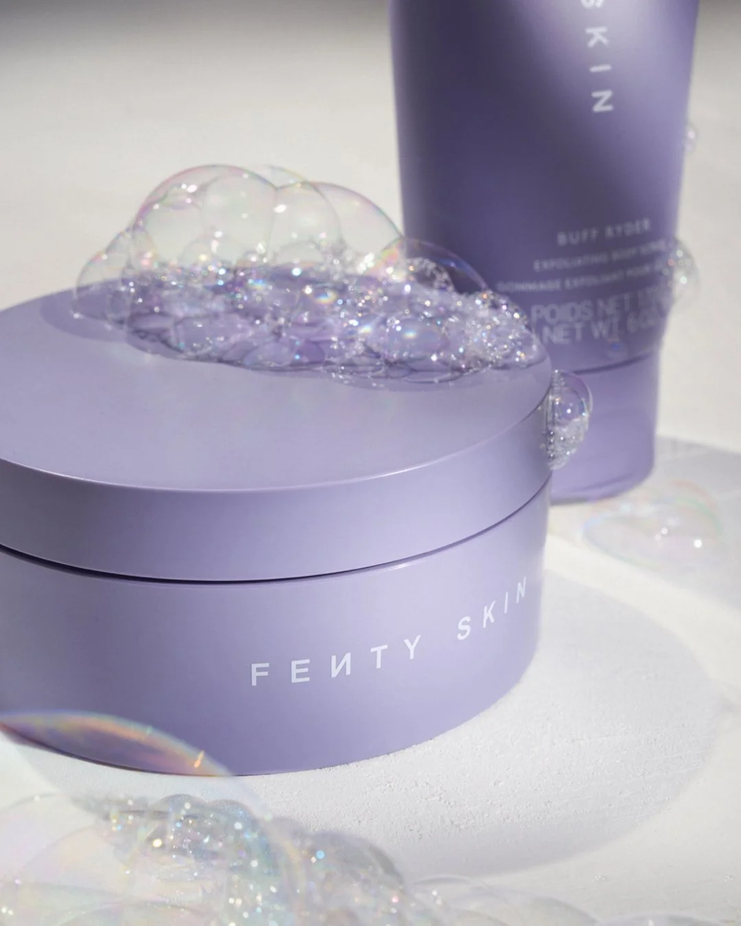 A purple Fenty Skin container with soap bubbles on top, a purple Fenty Skin bottle in the background, and iridescent bubbles on a white surface.
