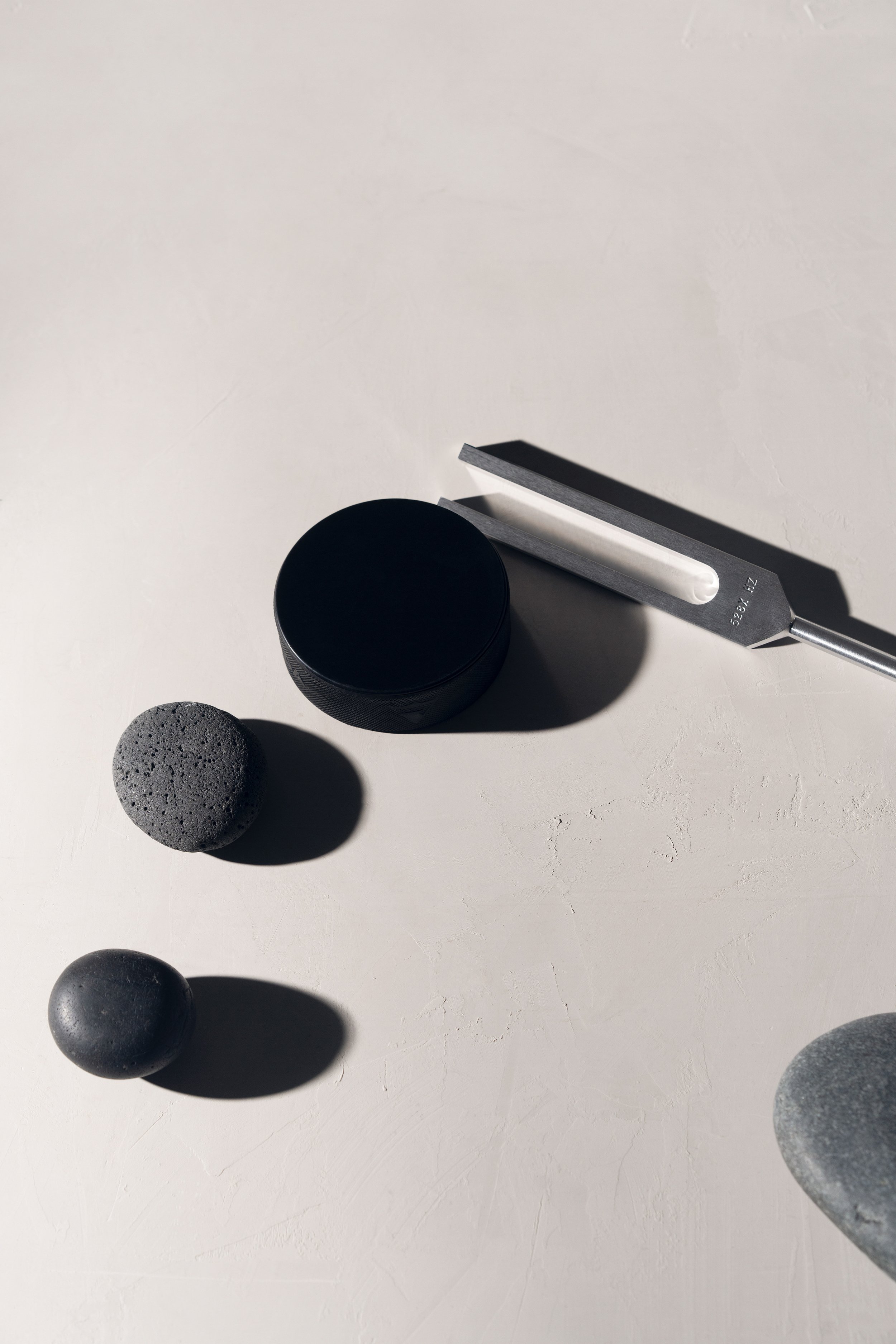 Minimalist arrangement of objects on a white surface, including a  stone, a black puck, a stainless steel Biofield Tuning fork.