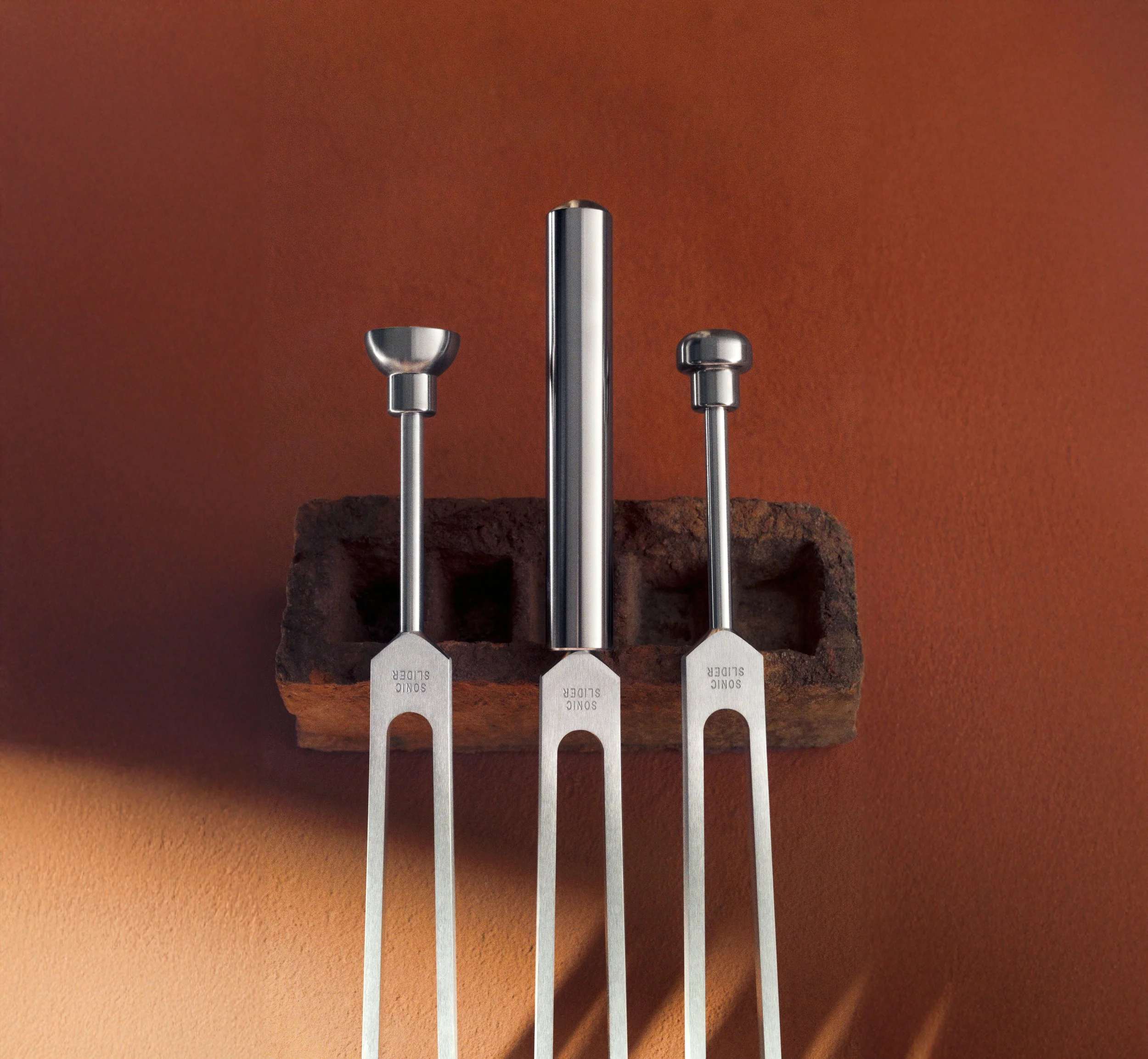 Three Biofield Tuning forks, resting on a brick wall against a brown textured wall.