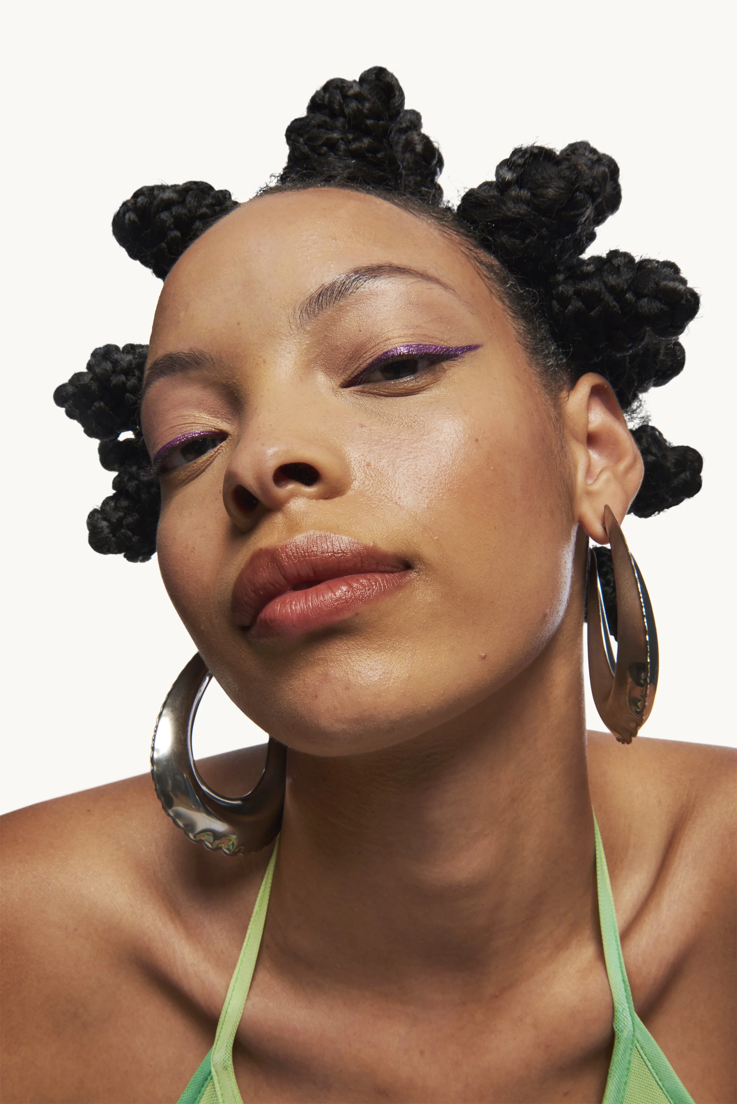 Close-up portrait of a woman with natural hair styled in bantu knots, wearing large silver hoop earrings and a green top, with subtle makeup and purple eyeliner.