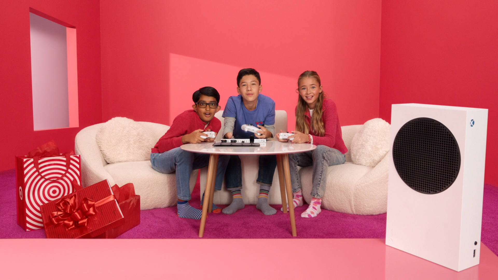 Three kids sitting on a white couch playing video games with controllers, set in a room with red walls, pink carpet, and a large white speaker on the right, with wrapped presents on the left.