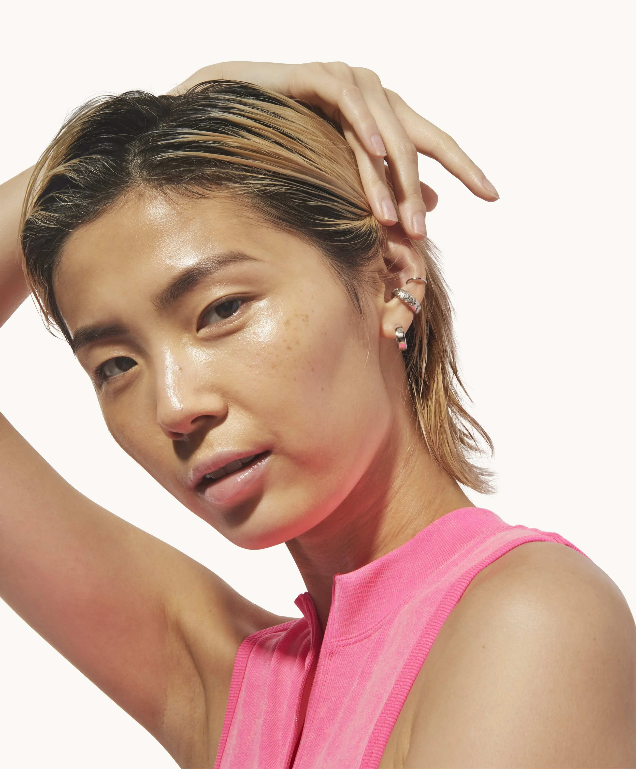 Close-up of a young woman with short, blonde and dark hair, wearing a pink sleeveless top, posing with her hand on her head against a plain white background.