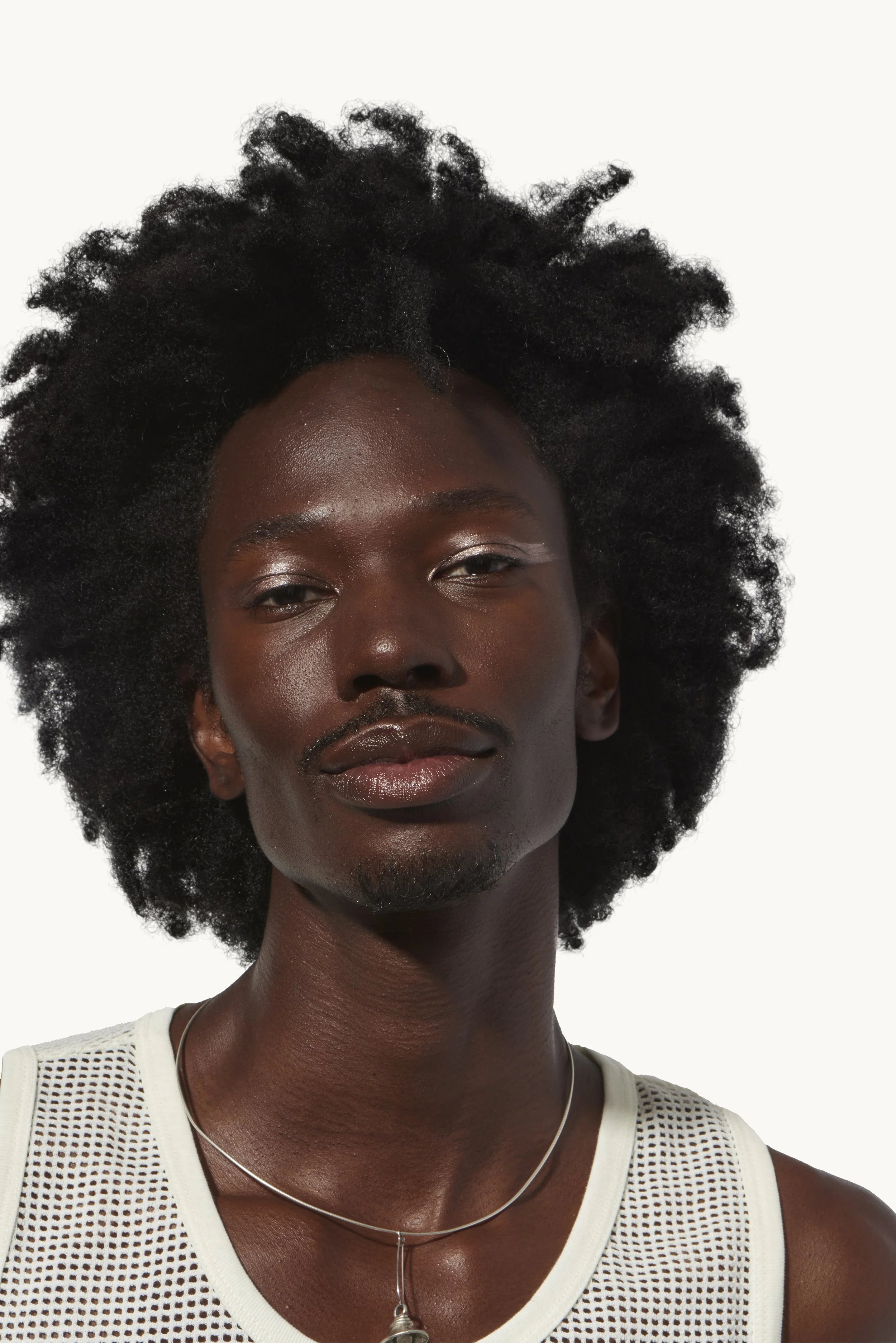 Close-up portrait of a young Black man with natural curly hair, wearing a white mesh tank top and a silver necklace, looking confidently at the camera against a plain white background.