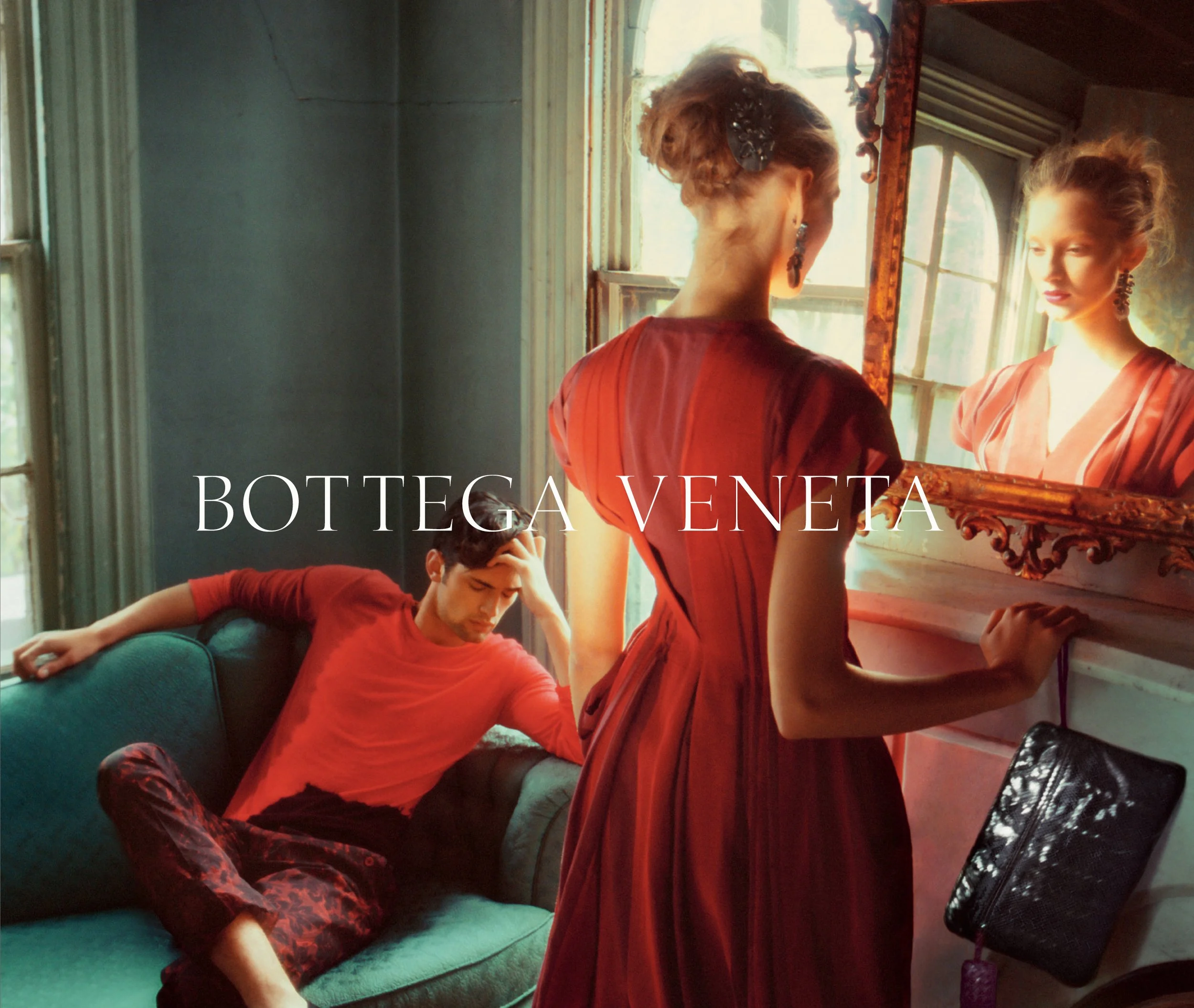 A young woman in a red dress looks into a mirror, with her back turned and a black hair accessory. Another woman in a matching dress stands by a window, and a young man in a red shirt and patterned pants sits on a teal couch nearby, resting his head on his hand. The room has large windows, wooden frames, and the Bottega Veneta logo overlaid.
