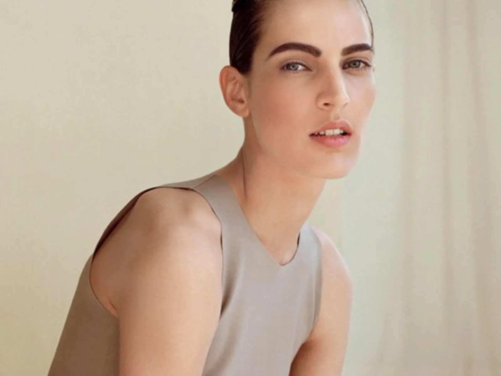 A woman with fair skin, dark brown hair, and blue eyes looks directly at the camera, wearing a sleeveless beige top, against a light-colored background.
