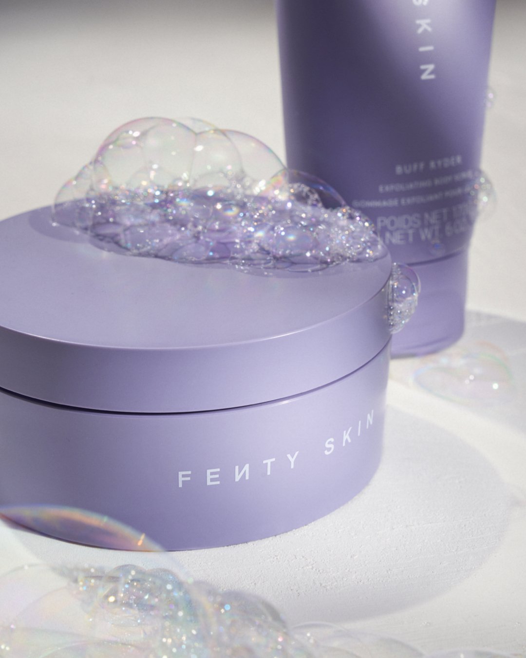 A purple Fenty Skin skincare product container with soap bubbles on top, alongside a matching purple bottle in the background, on a white surface.