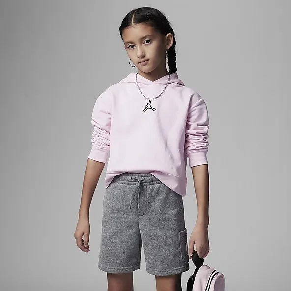 A girl wearing a pink hoodie with a small logo on the chest, gray shorts, and holding a small backpack, standing against a plain gray background.